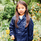 A young girl stands in a garden, wearing a navy Originals Waterproof Jacket, surrounded by flowers.